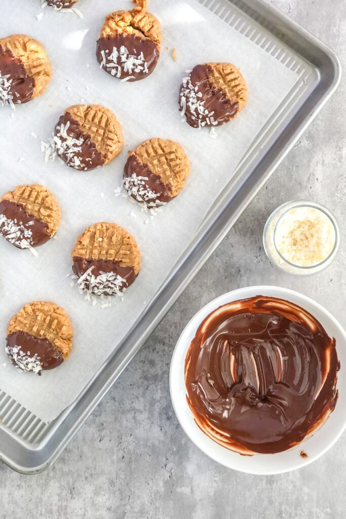 chocolate-dipped coconut peanut butter cookies on a baking sheet with a bowl of melted chocolate
