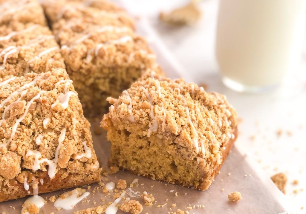 Biscoff spice crumb cake cut into squares with icing drizzle sitting on a wooden board with a glass of milk behind it